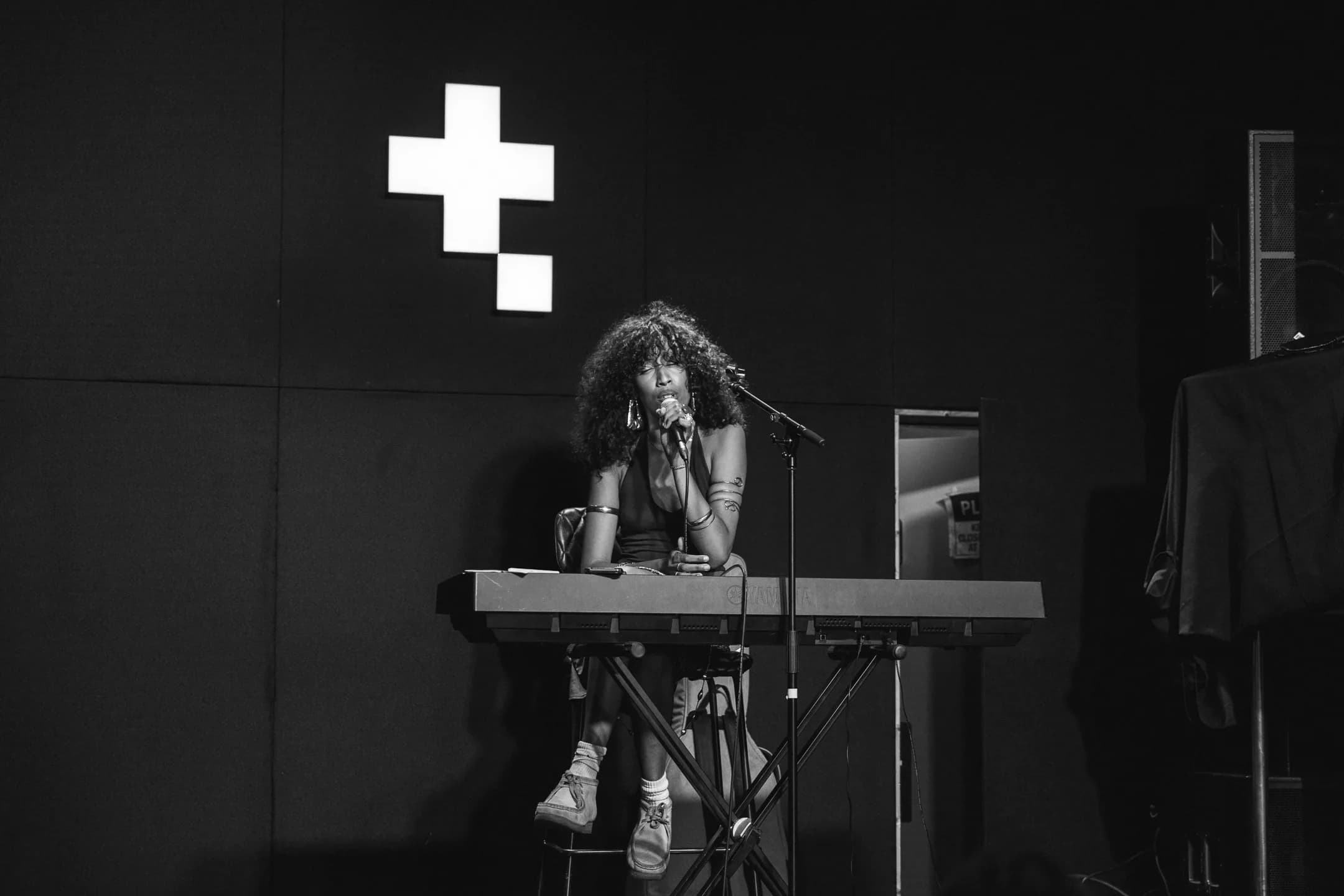 A live performer singing at a keyboard on stage at Two Tribes Campfire, the Two Tribes logo lit up behind her